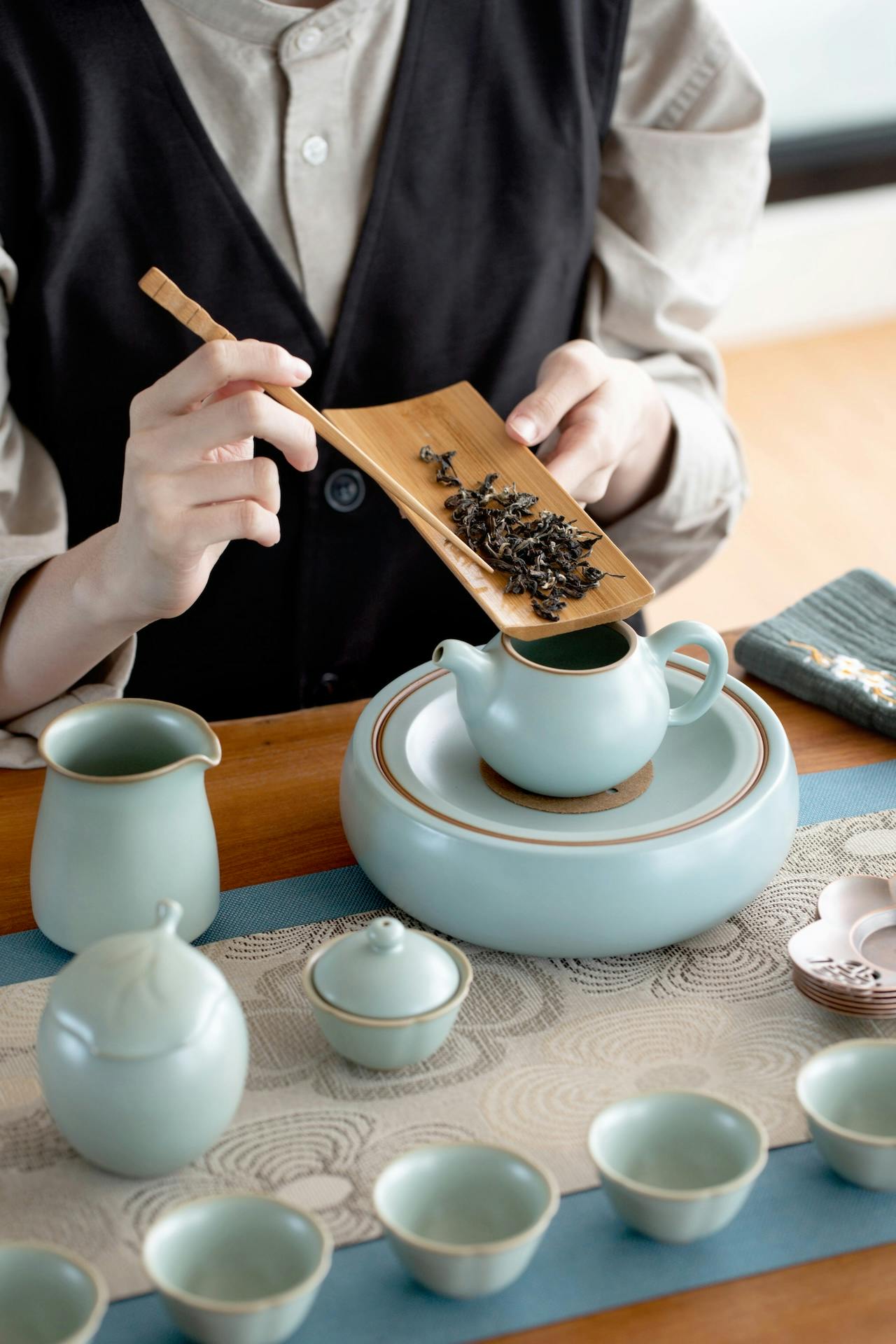 A person preparing tea using a bamboo scoop to add tea leaves into a teapot, surrounded by light blue ceramic teaware.