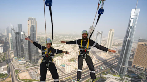 Two people in harnesses, arms outstretched, high above a cityscape with skyscrapers and highways.