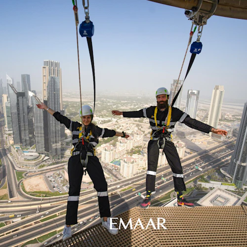 Two people in harnesses, arms outstretched, high above a cityscape with skyscrapers and highways.