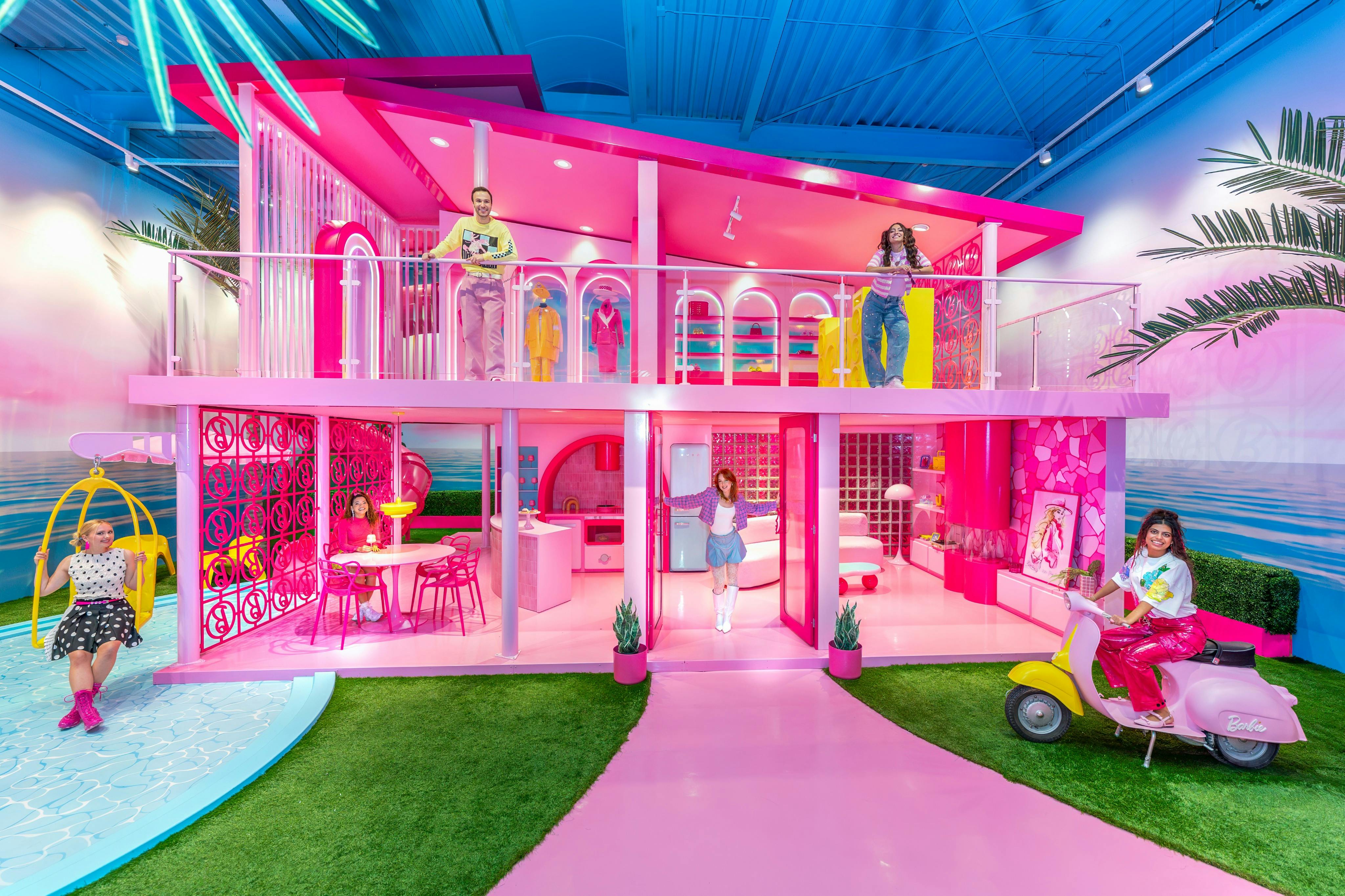 Four people pose in a two-level, life-sized pink dollhouse. One sits on a pink scooter, while others stand around inside and on the balcony.