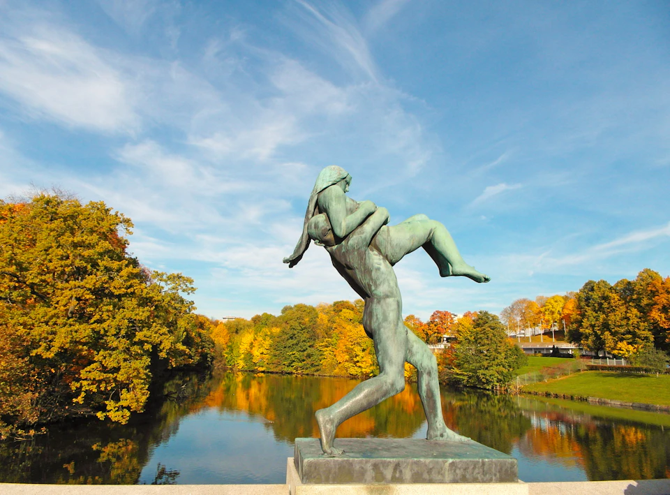 Bronze statue of a figure carrying another over a lake with autumn trees and a blue sky in the background.