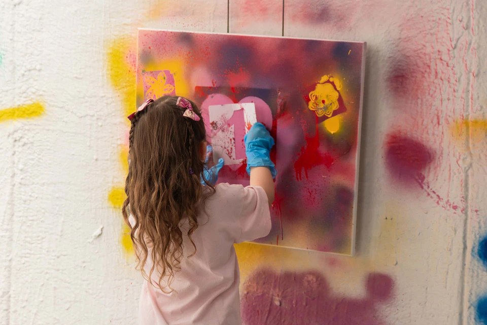 A child with long hair and blue gloves spray paints on a canvas with multicolored graffiti.