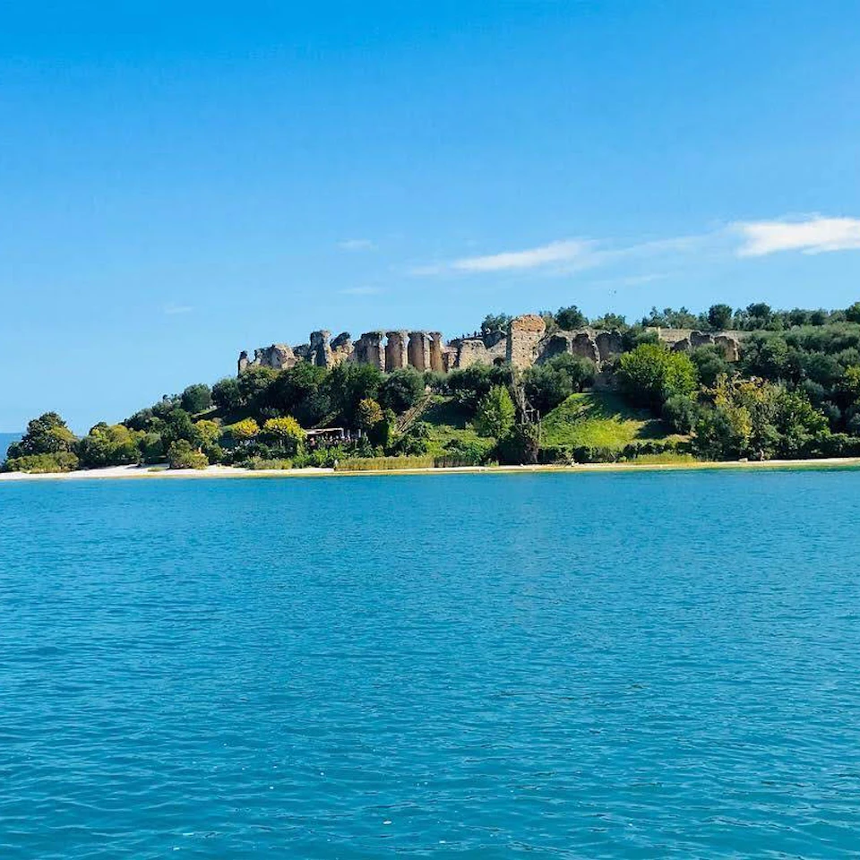 A historic stone structure surrounded by greenery on a hill, viewed across a calm body of water under a clear blue sky.