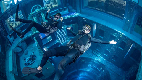 Two scuba divers float underwater in a large pool, with buildings and objects visible in the background.