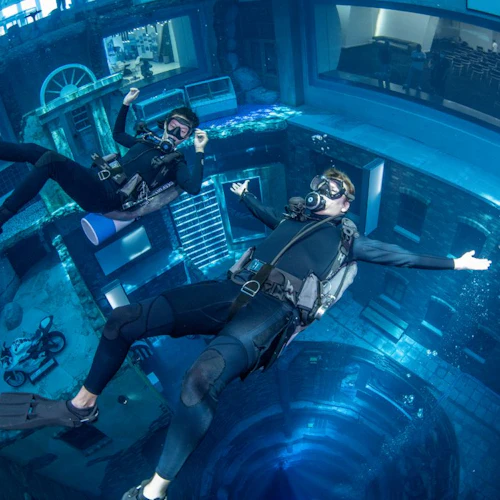 Two scuba divers float underwater in a large pool, with buildings and objects visible in the background.