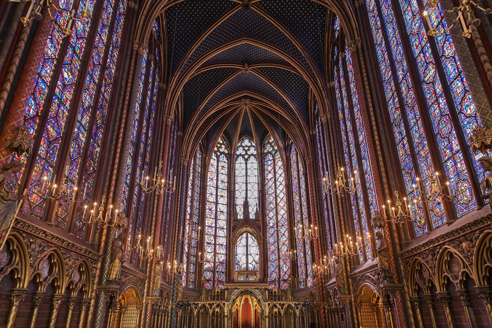 A richly decorated Gothic-style chapel interior with tall stained glass windows, vaulted ceiling, and ornate chandeliers.