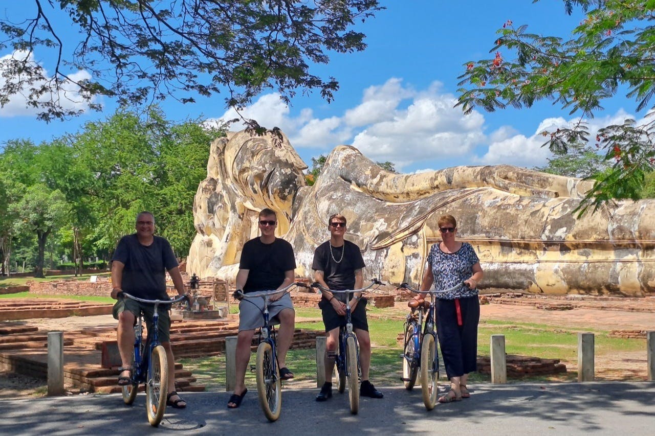 Quatro pessoas em bicicletas posando em frente a uma grande estátua de pedra reclinada num parque, num dia de sol.
