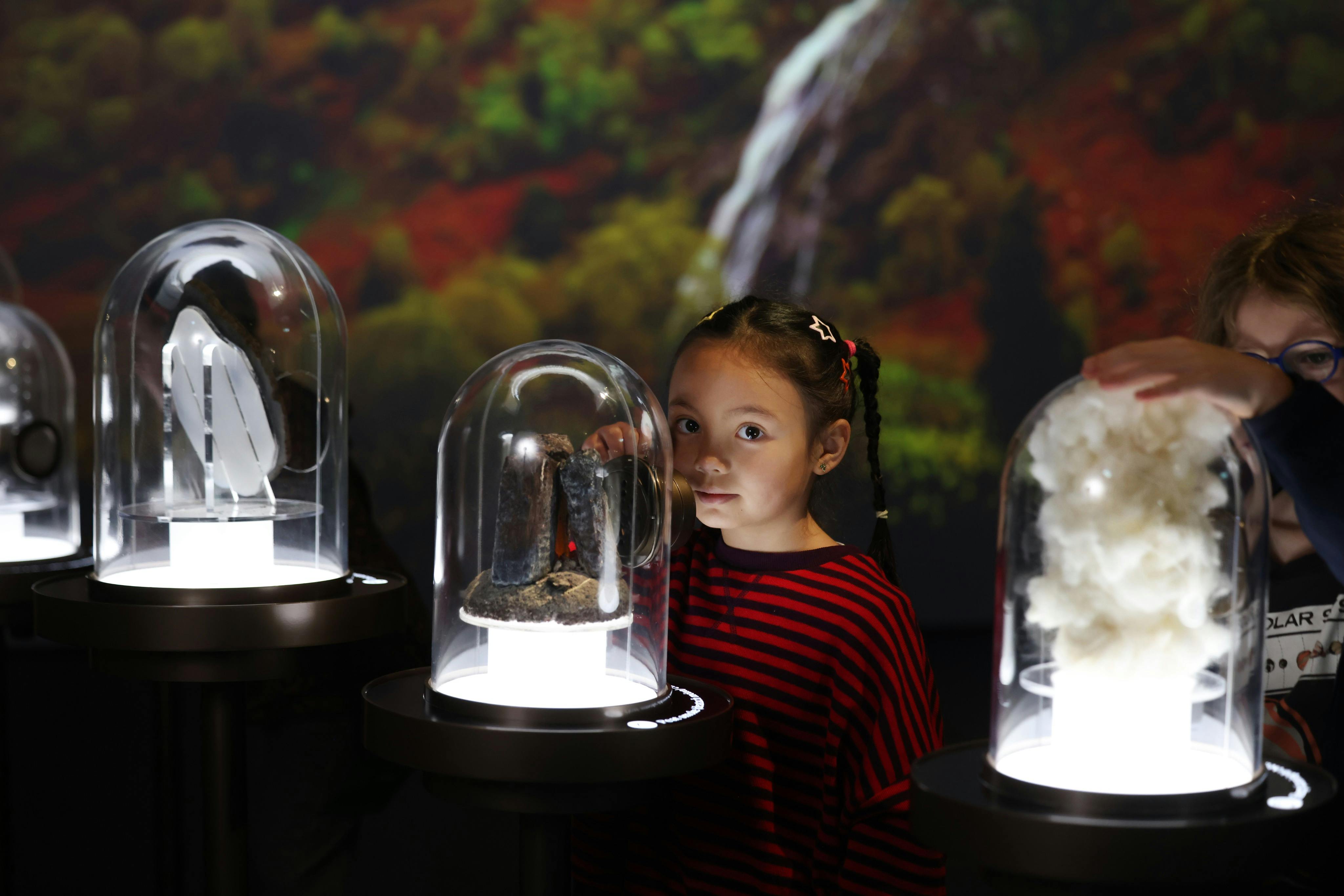 빨간 줄무늬 셔츠를 입은 어린 소녀가 유리 돔에 둘러싸인 물체를 바라보고 있으며, 그 뒤에는 자연을 주제로 한 배경이 있습니다.