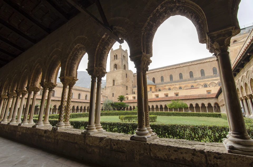 A medieval cloister with a view of an arched colonnade, a central garden, and a stone tower in the background.