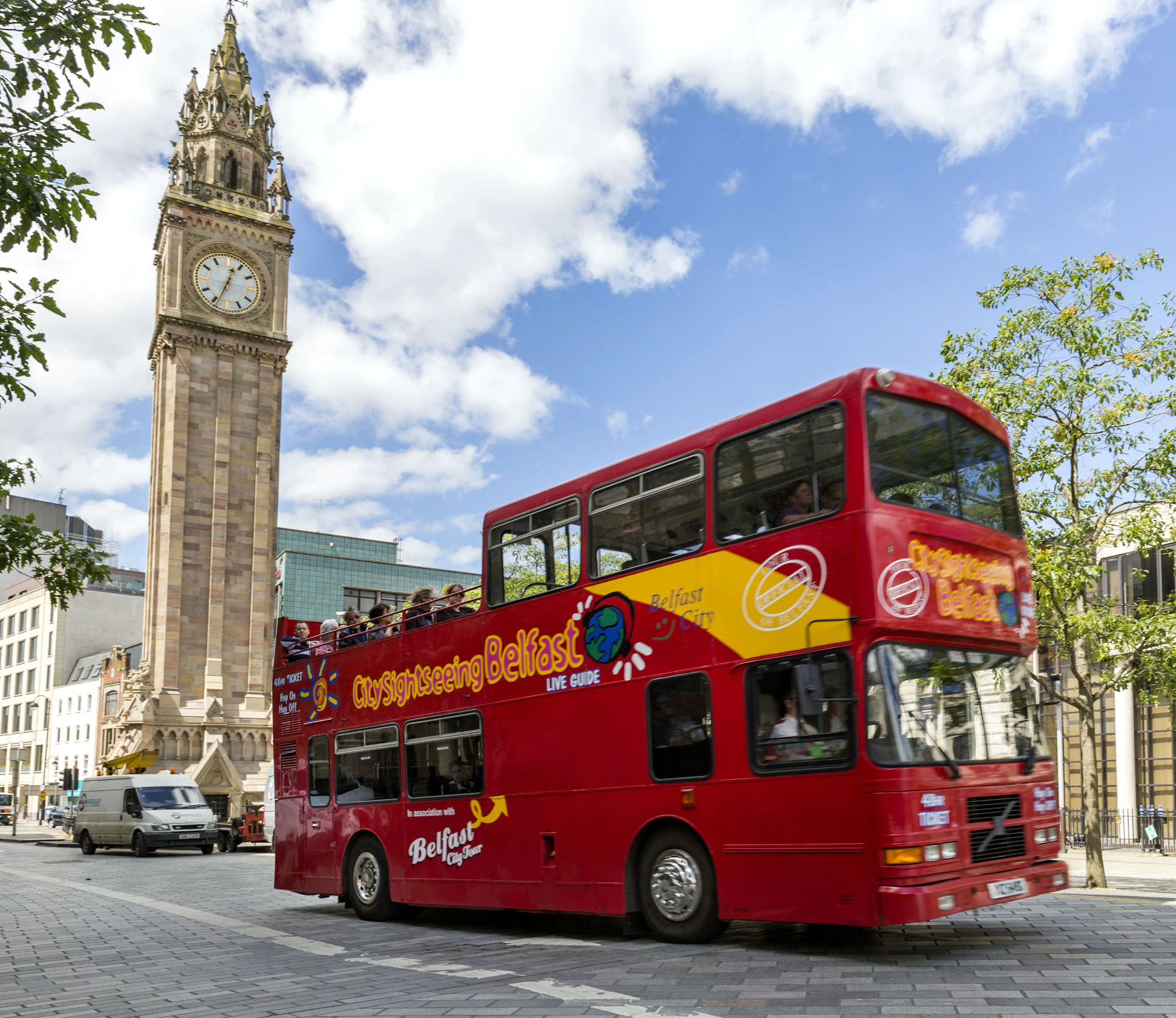 A red double-decker sightseeing bus in Belfast passes by a tall clock tower on a sunny day with scattered clouds.