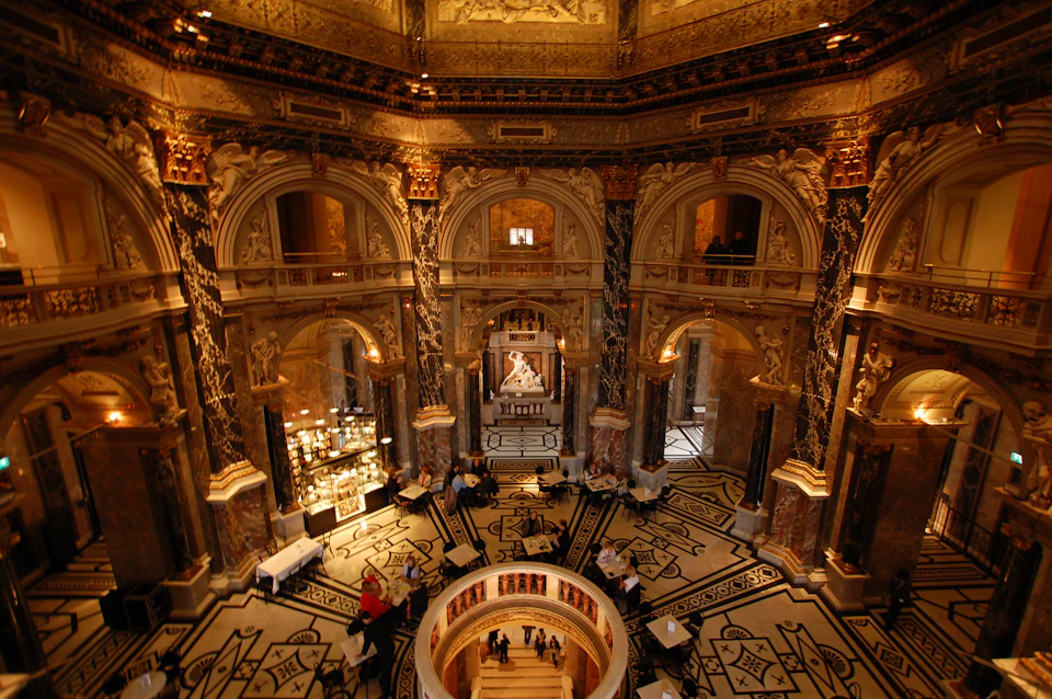 Ornate interior of a grand hall with marble columns, intricate carvings, and a central staircase. People are exploring and sitting.