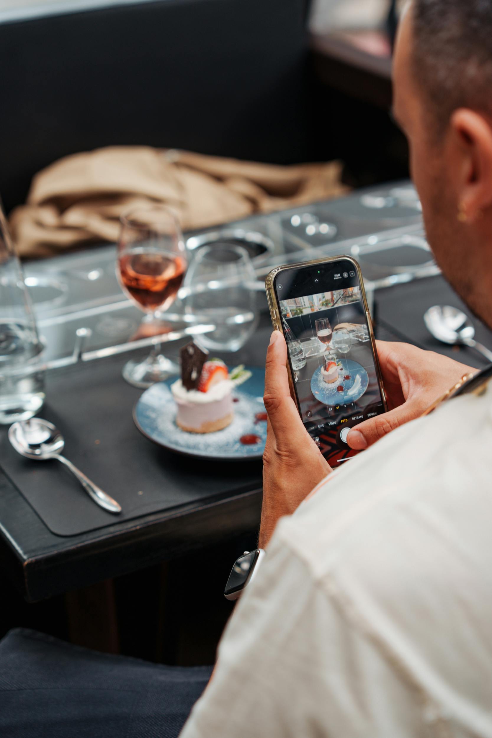 Iemand maakt met een smartphone een foto van een dessert op een blauw bord aan een gedekte tafel met glazen, bestek en een wijnglas.