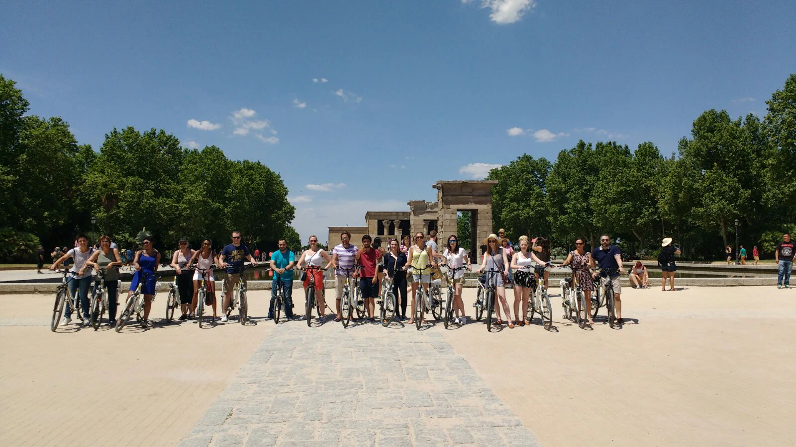 A group of people stand with bicycles on a stone path, with an ancient structure and trees visible in the background under a clear sky.