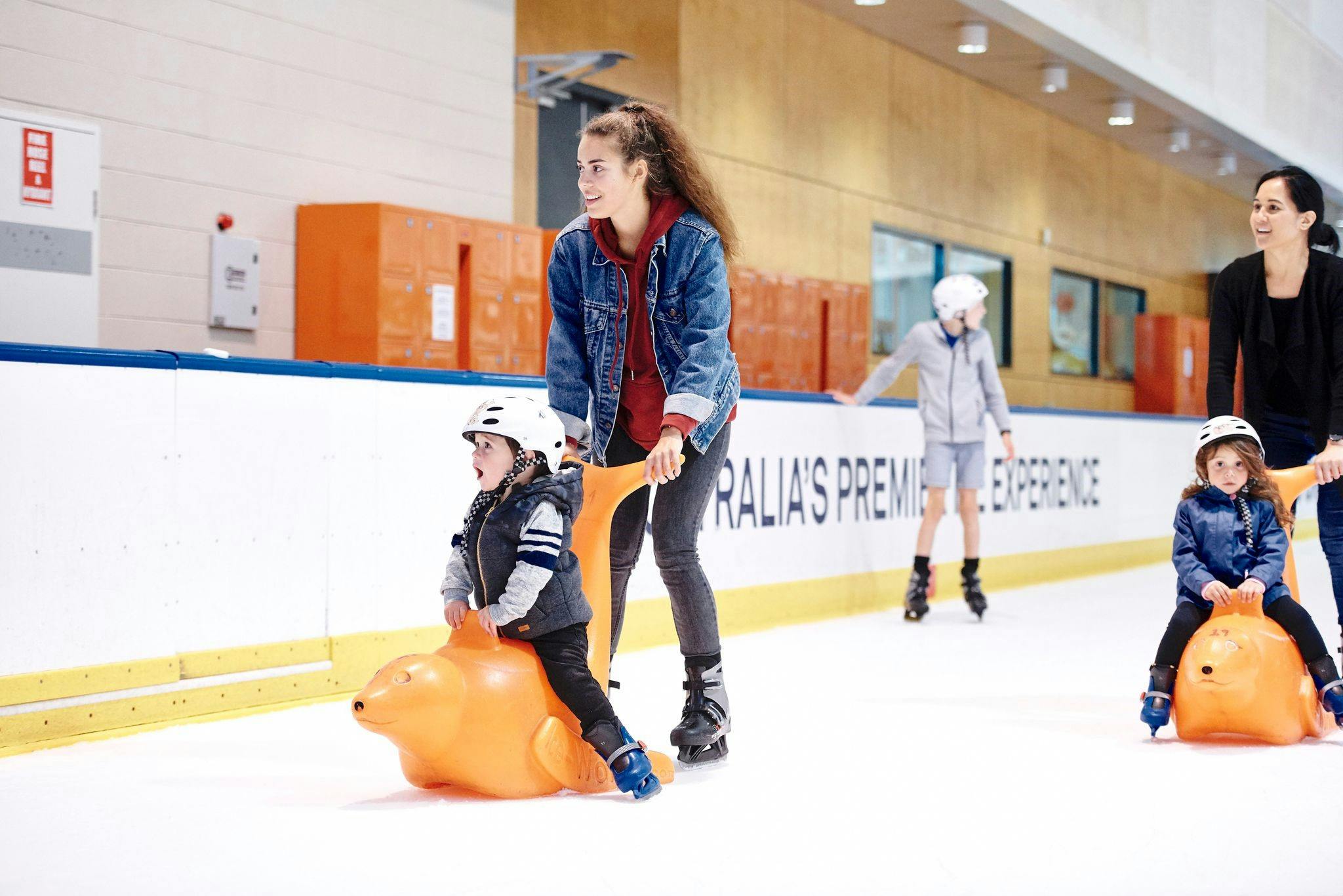 Two children sit on orange seal-shaped skating aids, each pushed by an adult, on an indoor ice rink with lockers in the background.