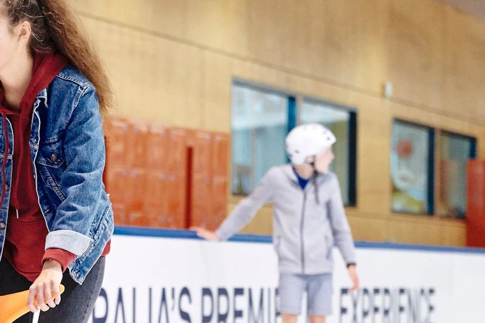 Adults and children ice skating in a rink, with children sitting on orange skating aids.