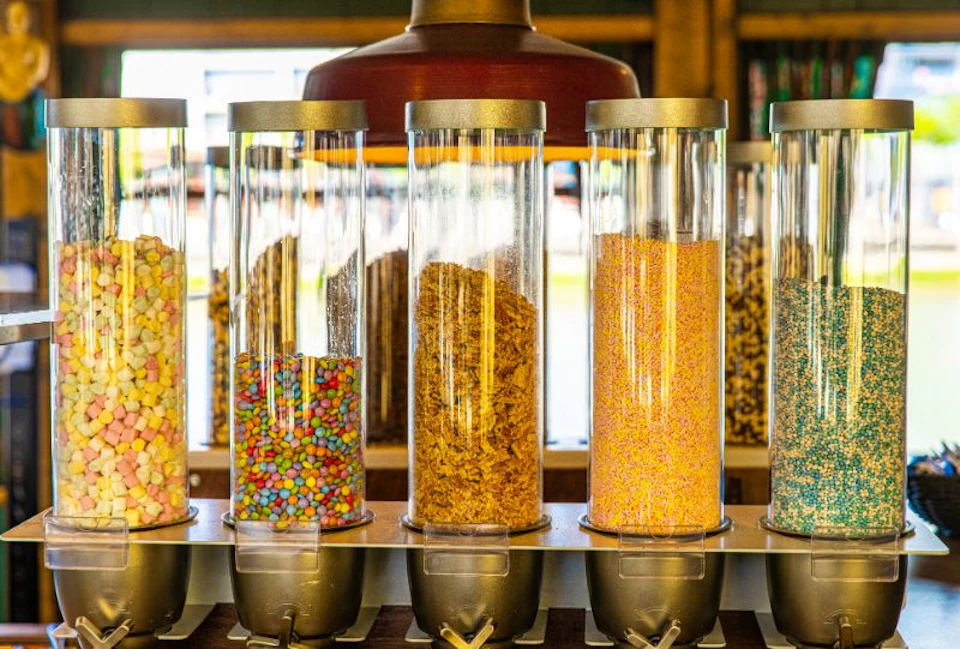 Transparent dispensers filled with various cereals and colorful toppings, each equipped with a silver handle at the bottom.