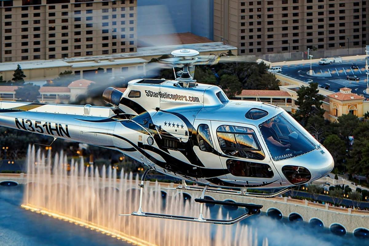 A helicopter with "5 Star Helicopters" branding flying near a fountain in front of buildings in a city.