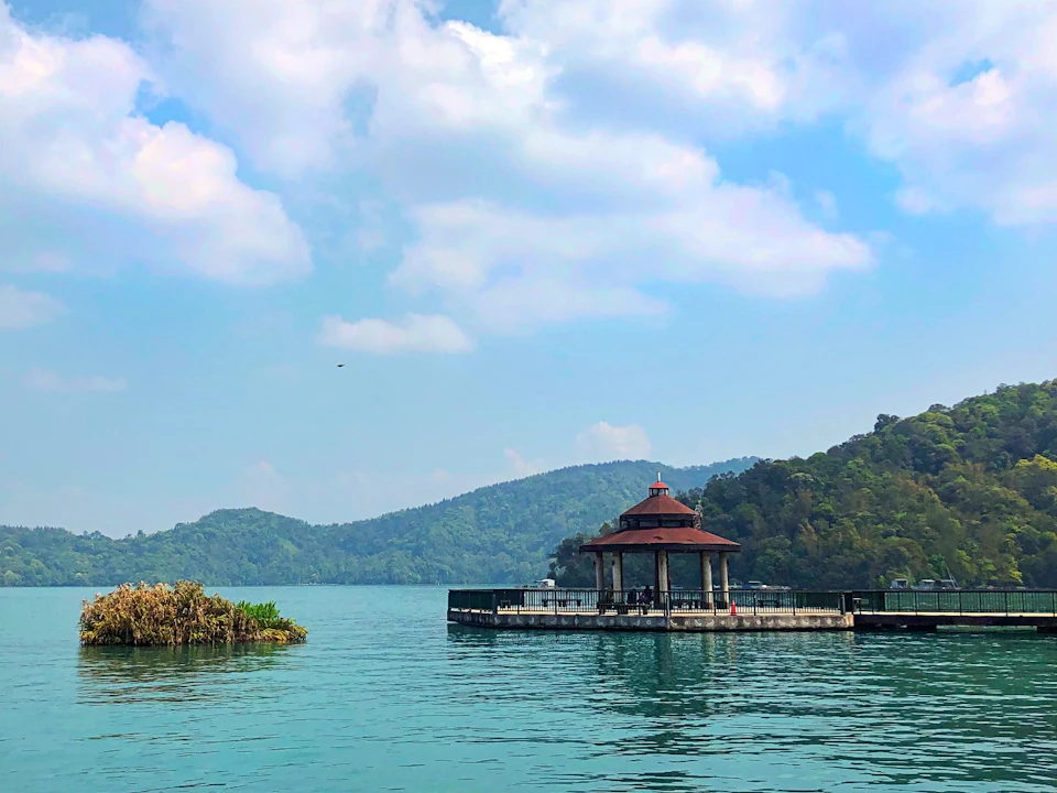 A gazebo on a platform in the middle of a lake, surrounded by hills under a partly cloudy sky.