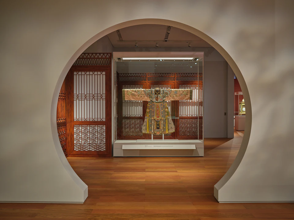 A traditional garment displayed in a glass case within a museum, viewed through a circular doorway and surrounded by wooden lattice walls.