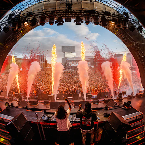 DJ: ar på en scen som uppträder för en stor folkmassa, med flammor och rökeffekter som skjuter upp, under ett välvt tak.