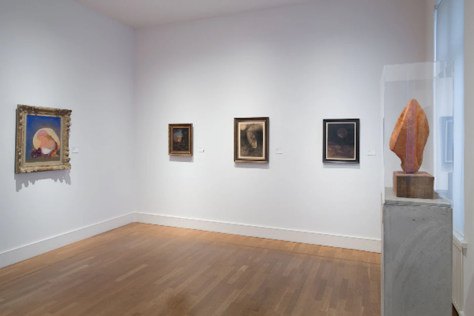 Three framed paintings on a white wall in a gallery with a wooden floor; a person's arm is partially visible on the right.