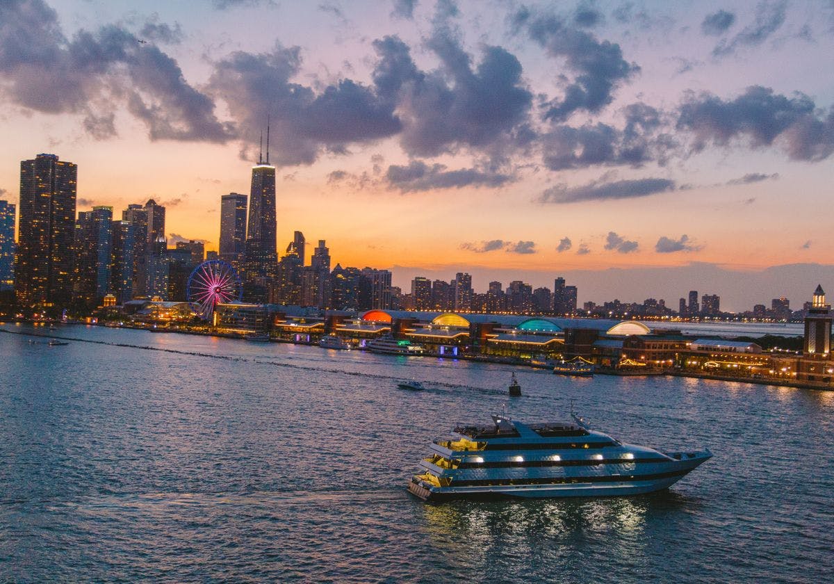 A boat cruises on a river at sunset with a city's skyline, featuring a Ferris wheel and illuminated buildings, in the background.