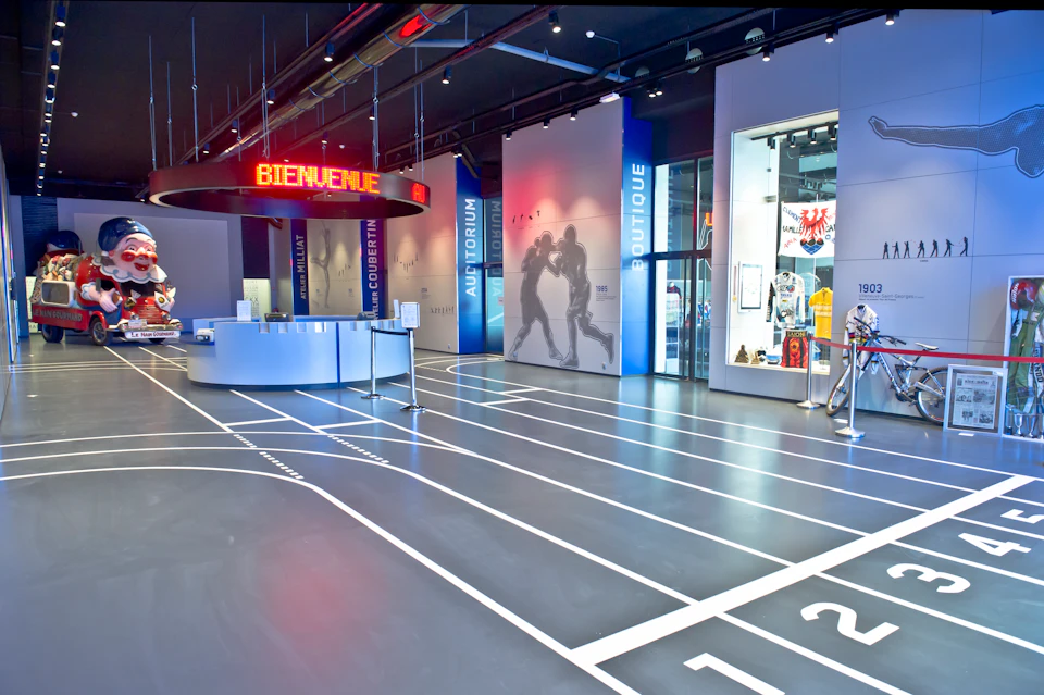 Modern exhibition hall with a track-themed floor, digital welcome sign, and sections labeled Auditorium, Boutique, and Coubertin.