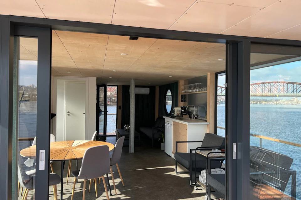 Interior of a modern houseboat with a dining area, kitchenette, and seating, featuring a view of water and a bridge outside.