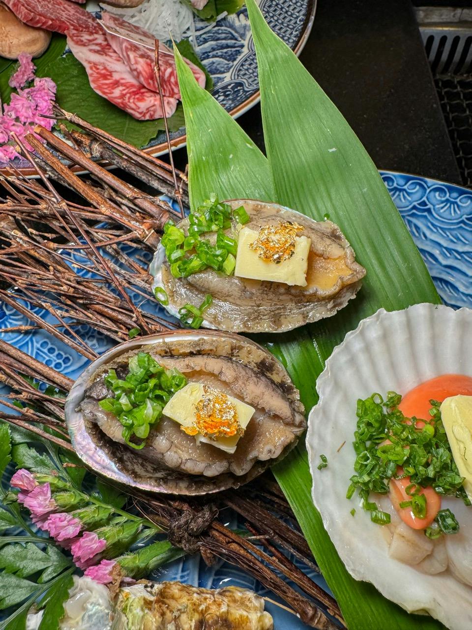 Two abalone shells with garnishes and butter are presented on green leaves, surrounded by decorative elements.