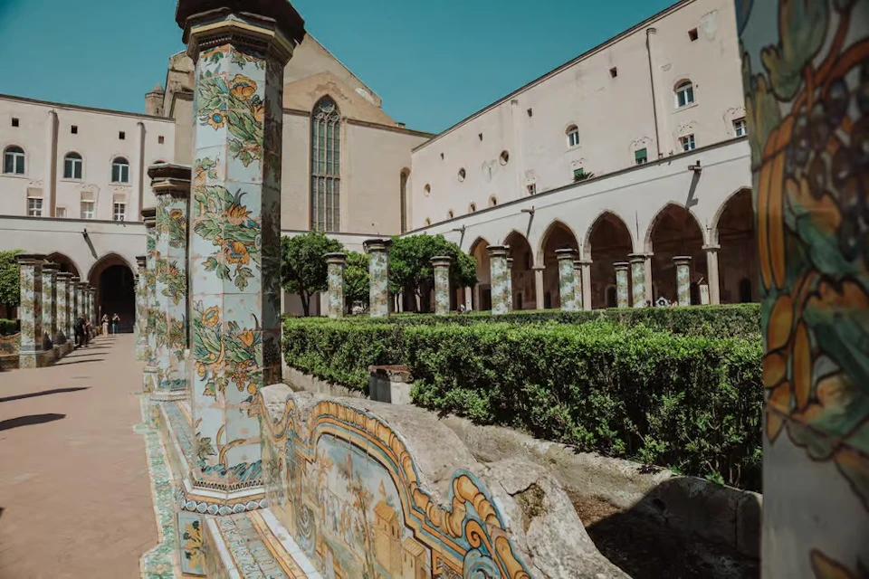 A cloister with painted ceramic columns, a manicured garden, and an old building featuring arches and windows under a clear sky.