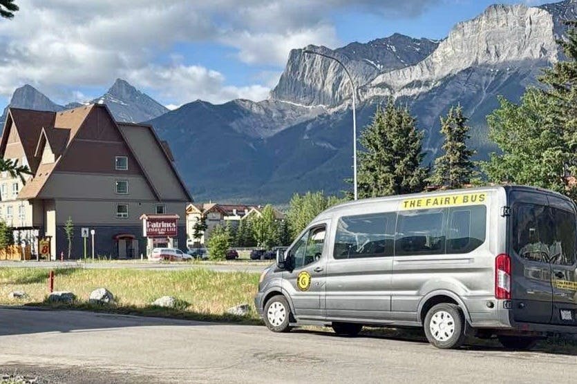 A gray van labeled "The Fairy Bus" is parked near a building with mountains in the background under a partly cloudy sky.
