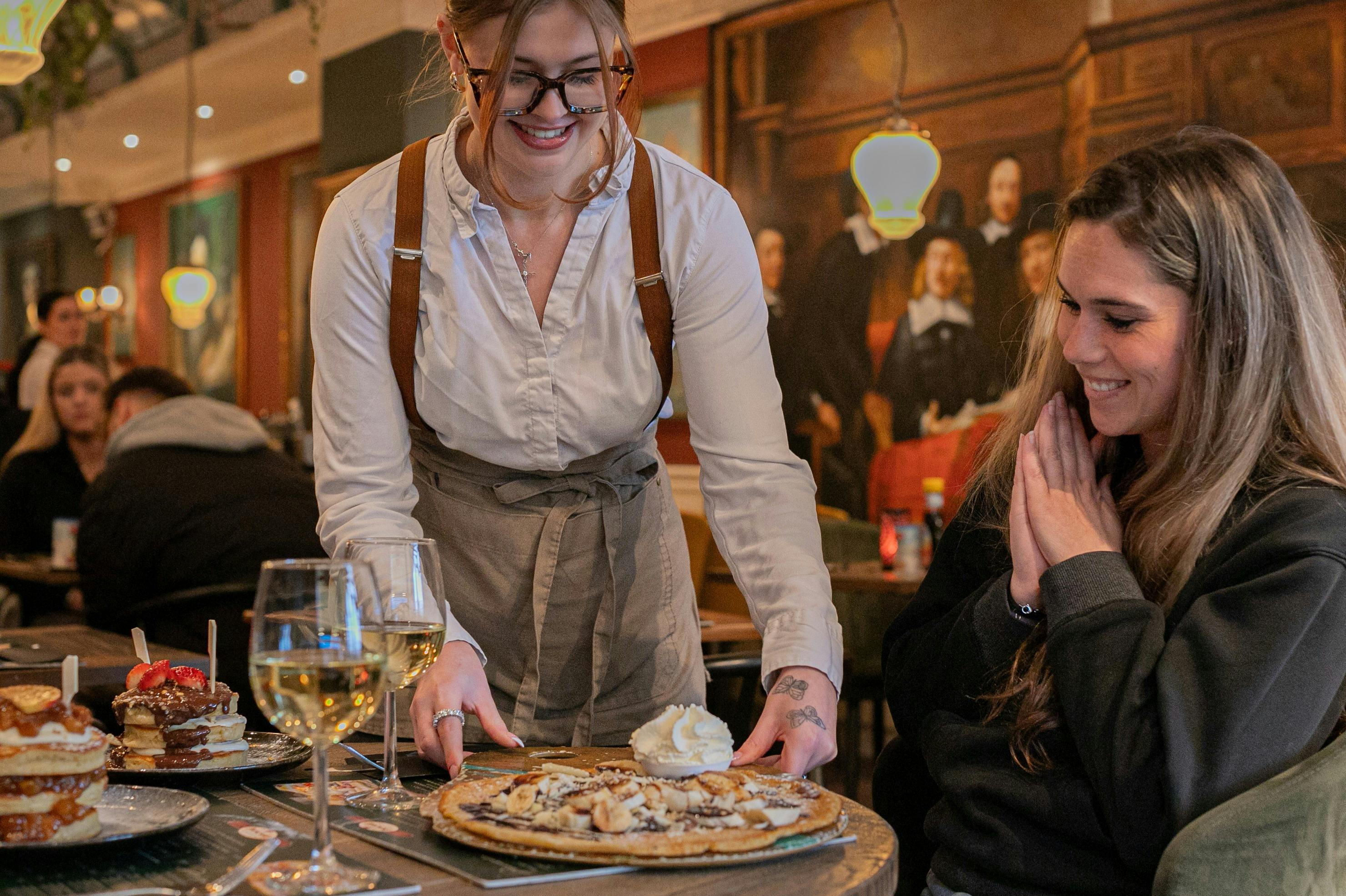 笑顔のサーバーは、暖かく明るいレストランで、喜んで座っている女性の前にホイップクリーム入りのデザートを置きます。