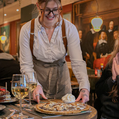 En leende server placerar en efterrätt med vispad grädde framför en glad sittande kvinna i en varmt upplyst restaurang.
