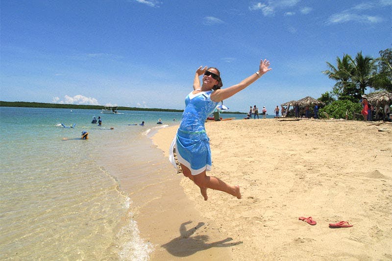 Woman in a blue dress jumps joyfully on a sandy beach, with people, huts, and a clear blue sky in the background.