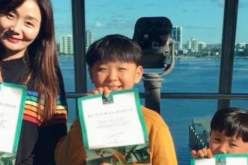 A woman and two children outdoors by a waterfront, holding certificates, with a city skyline in the background.
