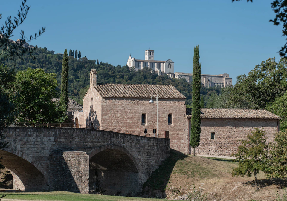 Stone buildings with a bridge, tall cypress trees, and a large hillside abbey in the background under a clear blue sky.