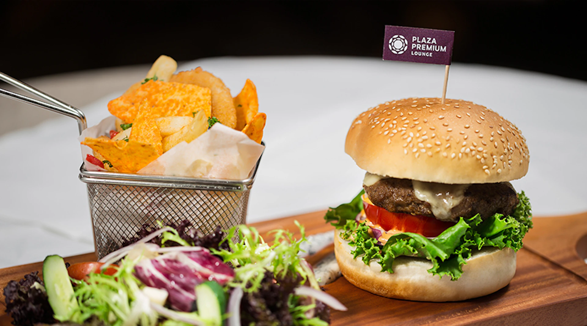 A cheeseburger with a lettuce salad and a side of fries in a basket, served on a wooden board with a branded flag.