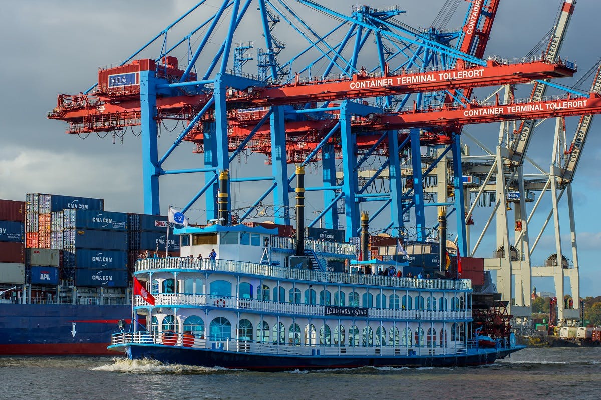 Paddelboot mit blauer Reling namens "Louisiana Star" auf dem Wasser, mit großen roten und blauen Kränen und Schiffscontainern im Hintergrund.