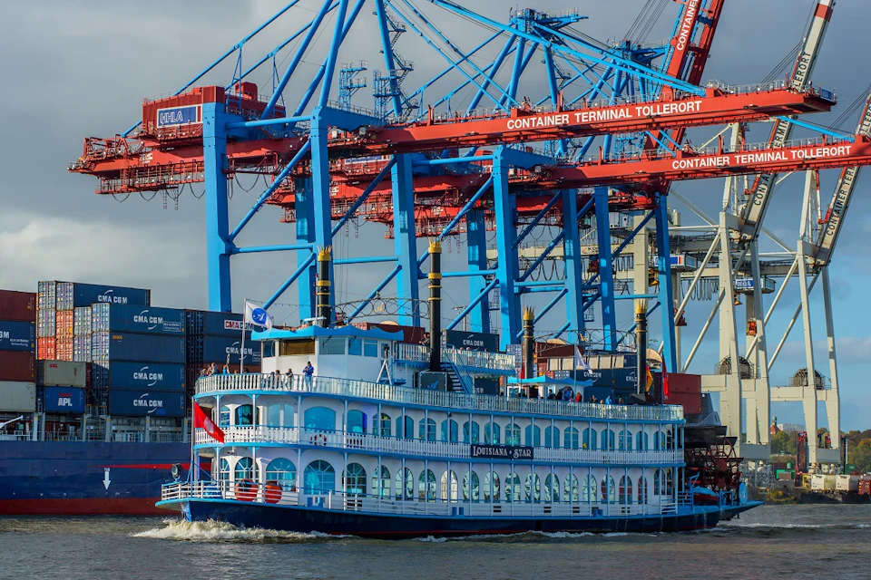 Paddle boat with blue railings named "Louisiana Star" on the water, with large red and blue cranes and shipping containers in the background.