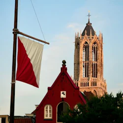 A tall church tower behind a red and white flag and a red building under a clear sky.