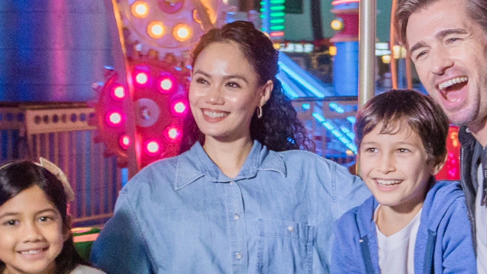 A family of four smiling while seated in a green amusement park ride, with colorful lights and structures in the background.