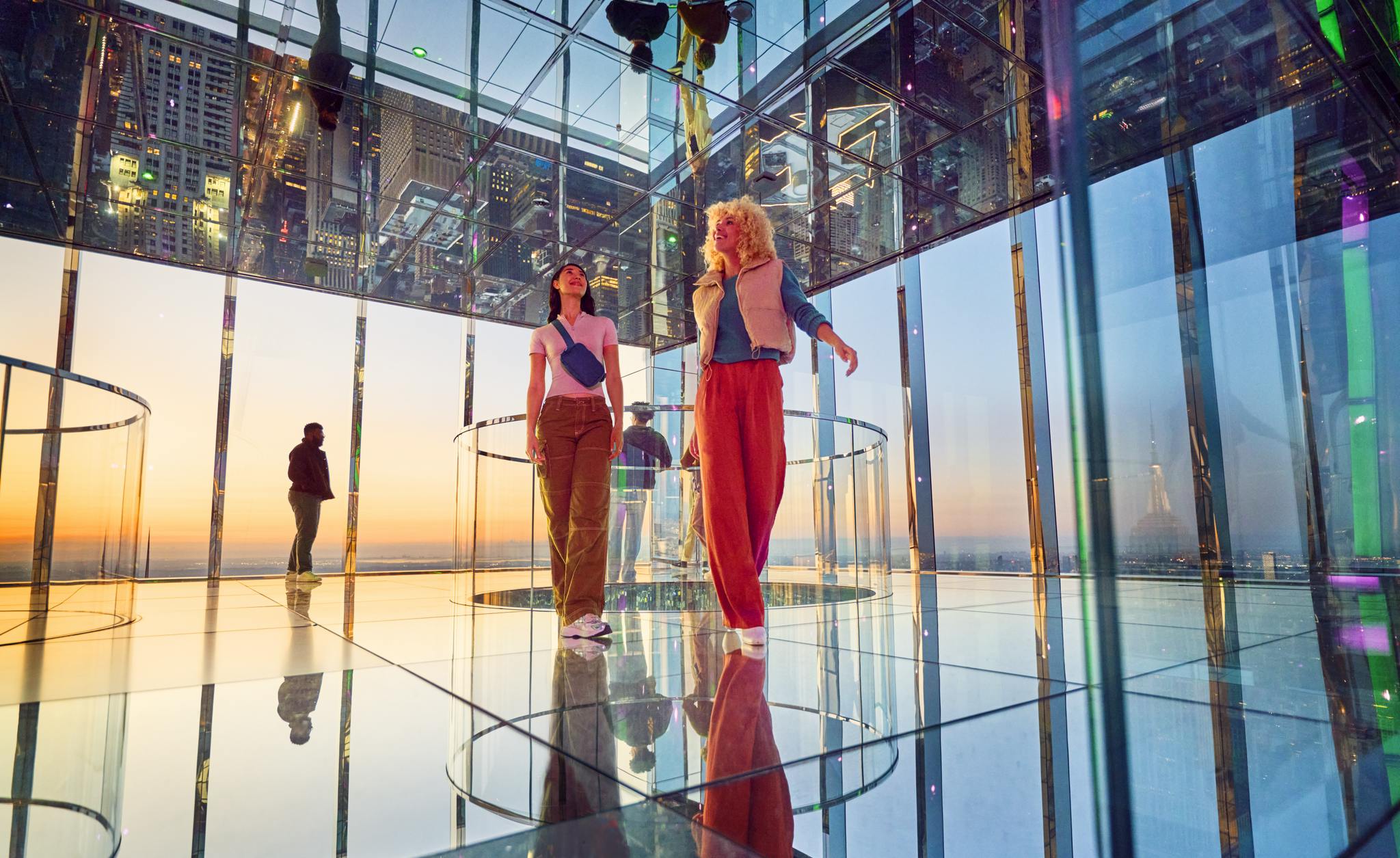 Dvě ženy stojí v zrcadlové místnosti s výhledem na město a barevnými odlesky při západu slunce. V pozadí pozoruje muž.