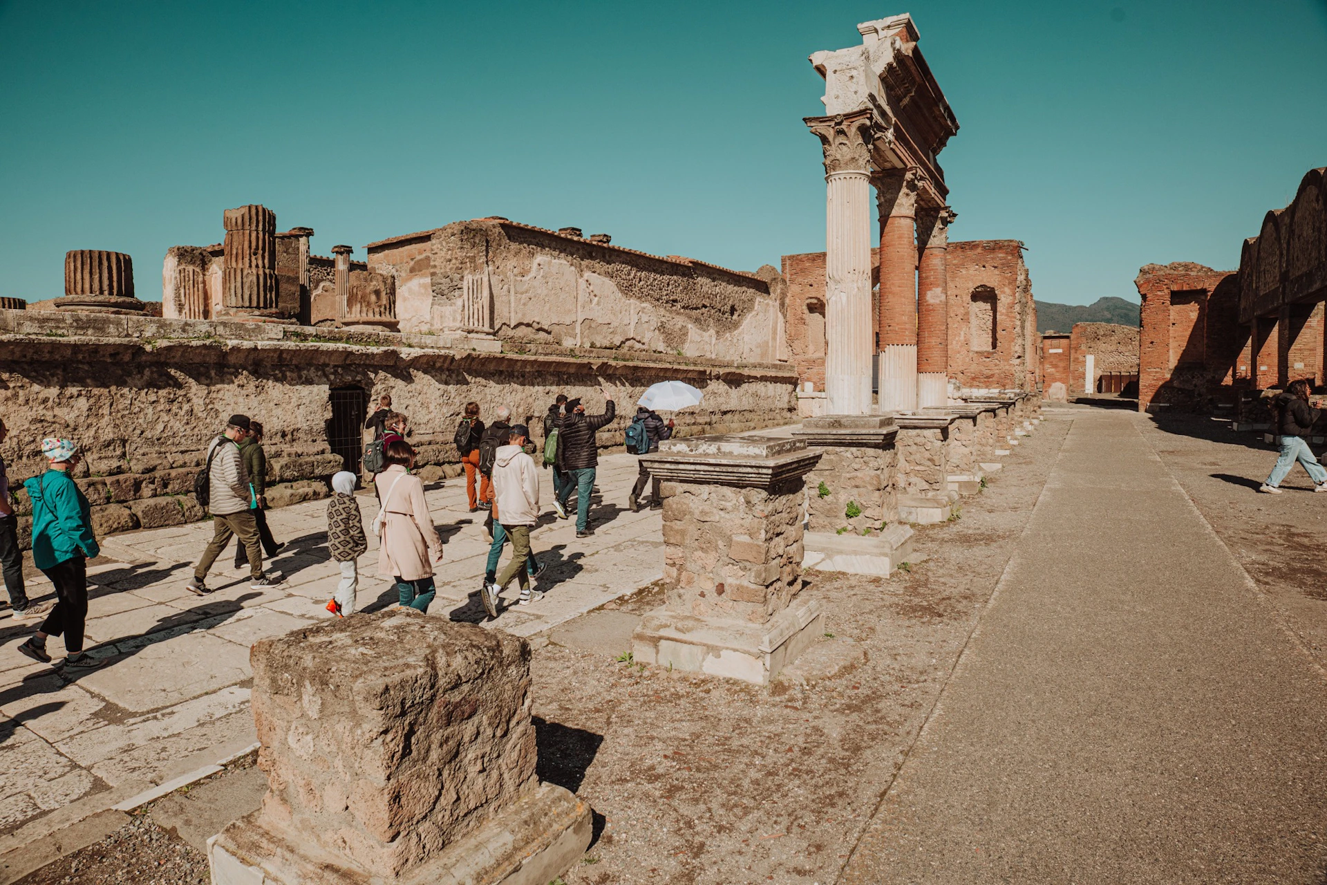 Tourists walk through ancient ruins with stone columns and bricks under a clear blue sky.