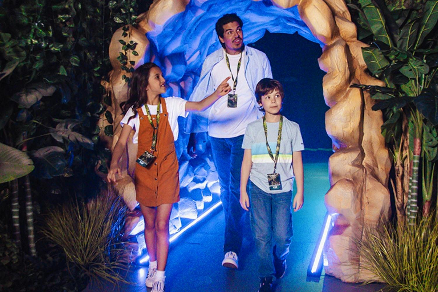 A man and two children walking through a cave-like, illuminated passageway with plants and rocks.