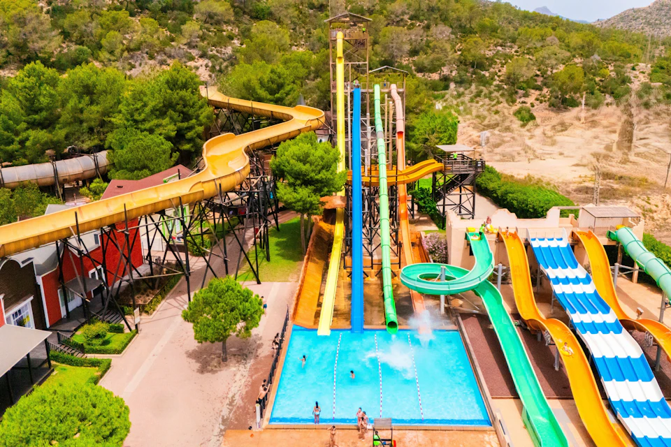 A water park features colorful water slides leading into a pool, surrounded by trees and a natural hillside.