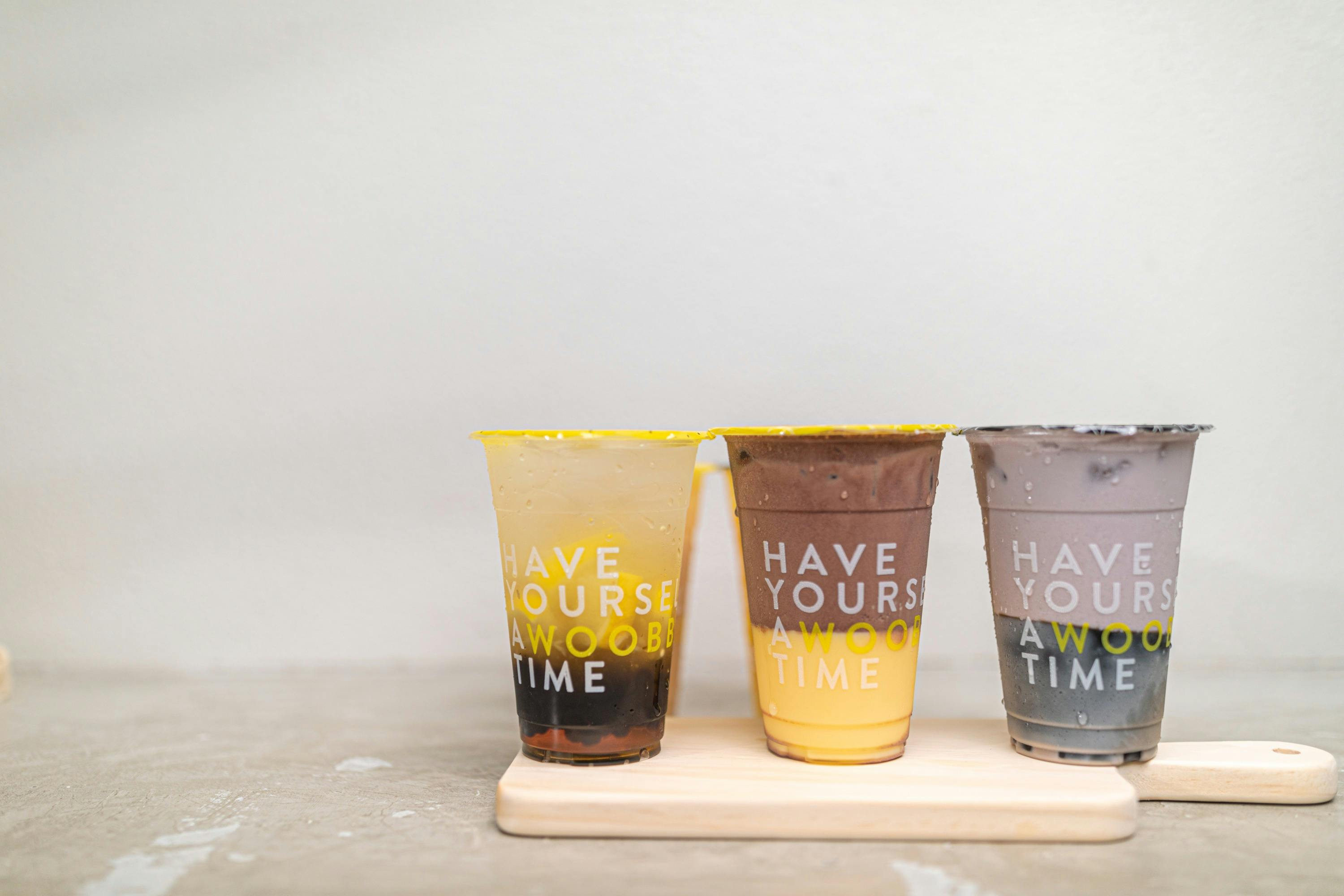 Three colorful drinks in transparent cups with yellow lids and text, placed on a wooden tray, against a plain background.
