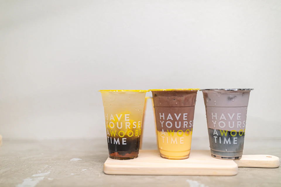Three beverages in plastic cups with yellow lids, placed on a light wooden tray, text on cups reads "HAVE YOURSELF A WOOBLY TIME."