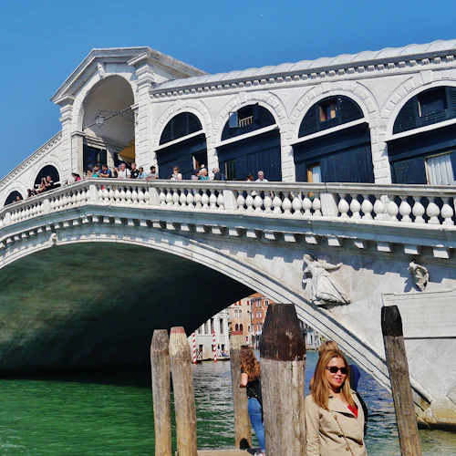 En historisk välvd bro över en kanal med båtar; en folkmassa på bron; byggnader och människor nära vattnet.