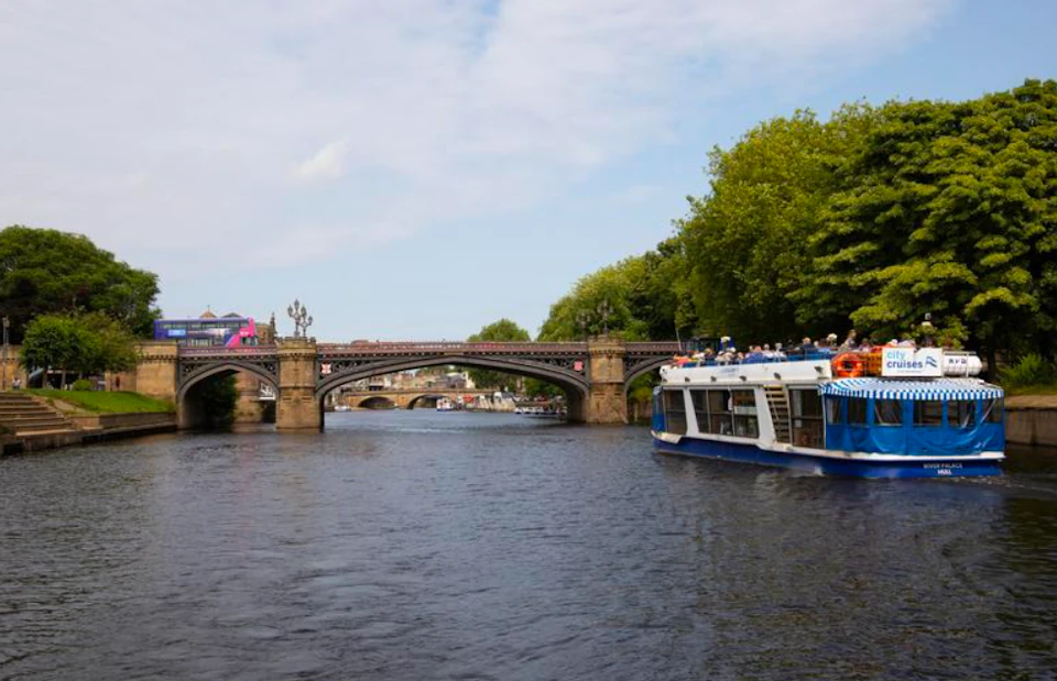 A river scene with a boat carrying passengers, a bridge in the background, and trees lining the riverbanks under a partly cloudy sky.