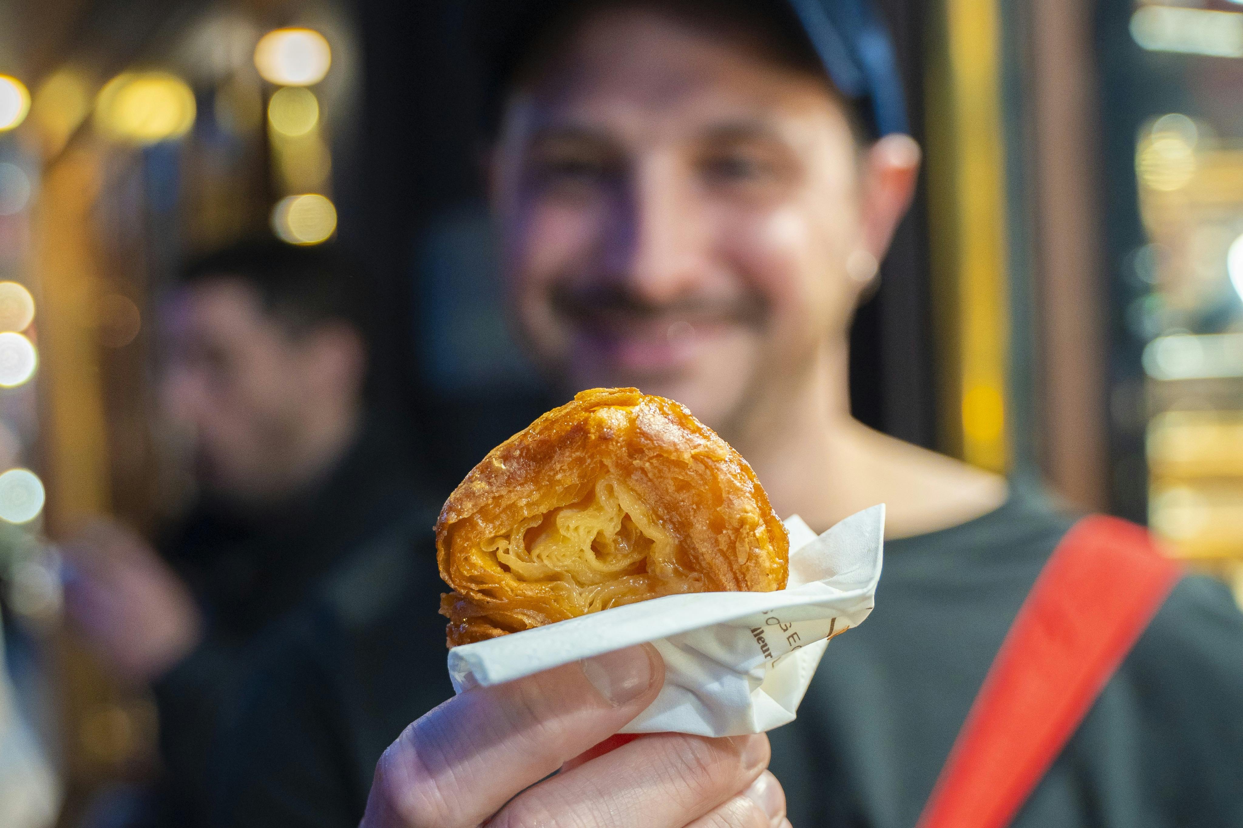웃고 있는 사람이 흐릿한 배경에 초점이 맞춰진 사진으로, 냅킨에 싸인 얇은 페이스트리를 들고 있습니다.